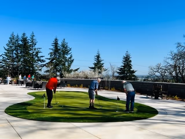 Three elderly men playing putting golf on a small green area outdoors at Boone Ridge Retirement Community, with trees and a clear blue sky in the background.