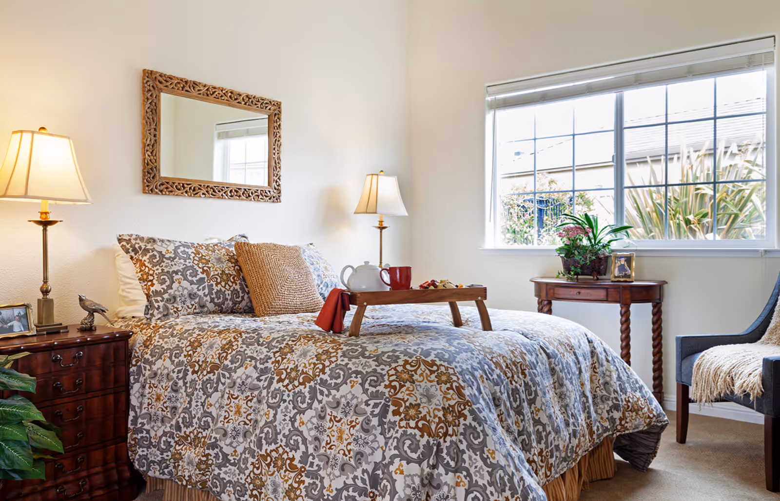 Decorated bedroom featuring a patterned bed with a breakfast tray, two bedside lamps, a mirror, and a window.