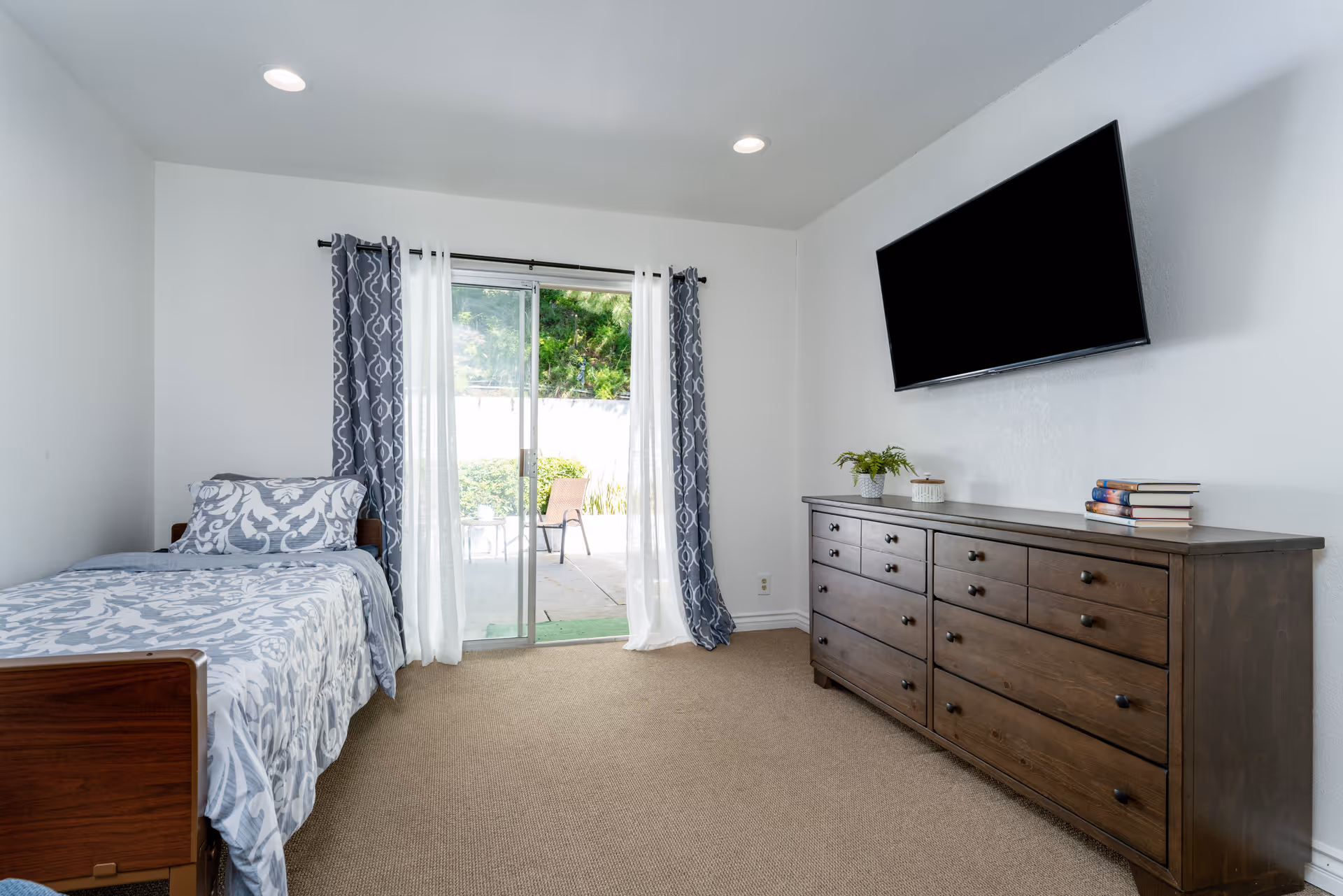 A bedroom with a single bed on the left side covered with a patterned gray and white bedspread and pillow. There is a sliding glass door with sheer white and patterned gray curtains leading to an outdoor patio. On the right side, there is a large wooden dresser with multiple drawers, a small plant, a candle, and a stack of books on top. Above the dresser, a flat-screen TV is mounted on the white wall. The room has beige carpet and recessed ceiling lights.