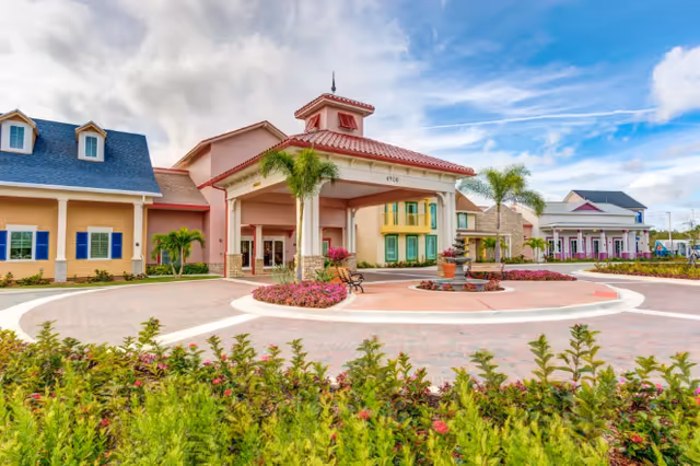 Front entrance of the Symphony at Stuart senior living building with a covered porte-cochere, circular driveway, palm trees, and landscaped gardens.