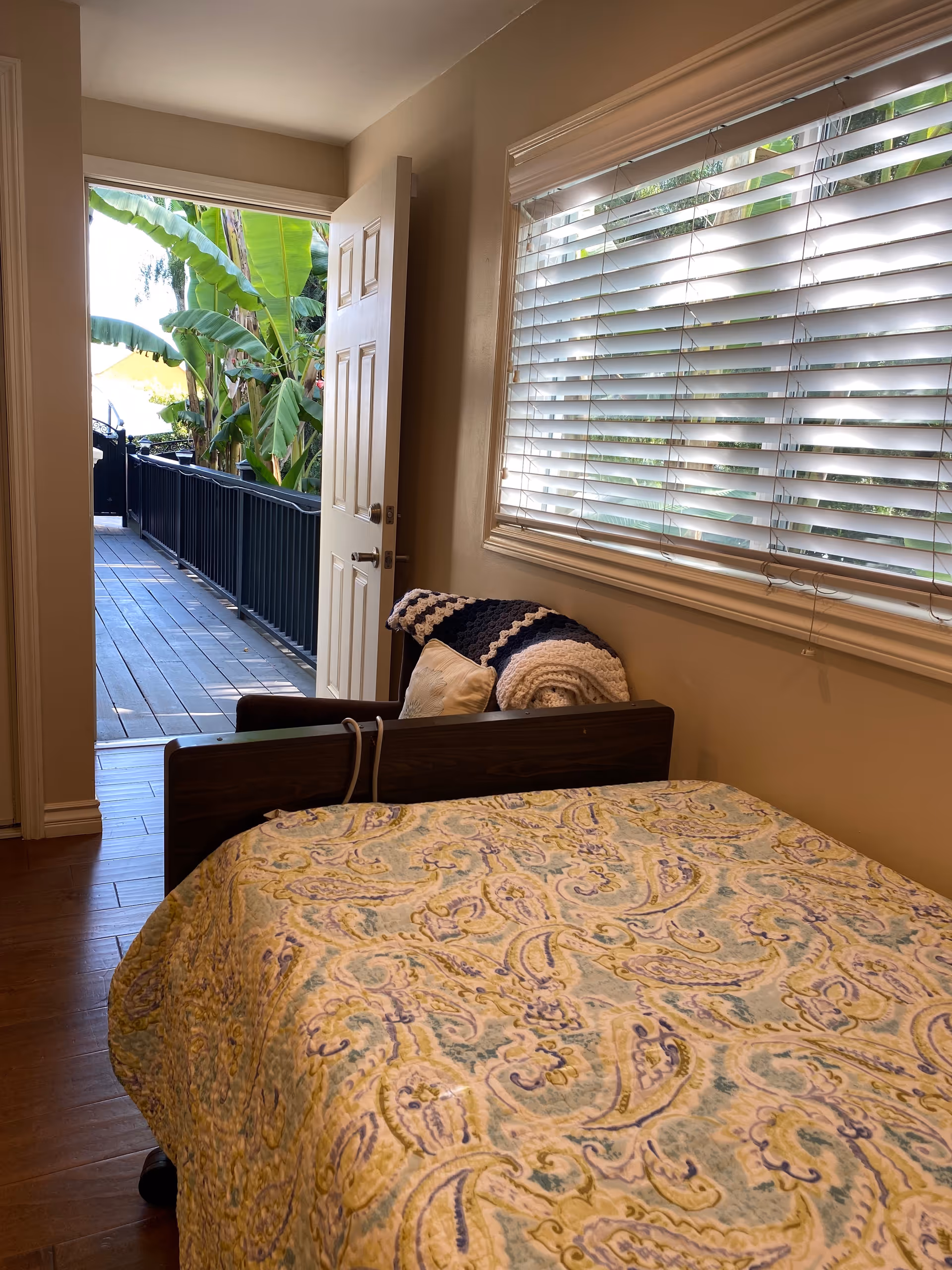 A bedroom with a bed covered in a patterned quilt, a chair with a crocheted blanket and pillow, a window with white blinds, and an open door leading to an outdoor walkway with tropical plants visible.