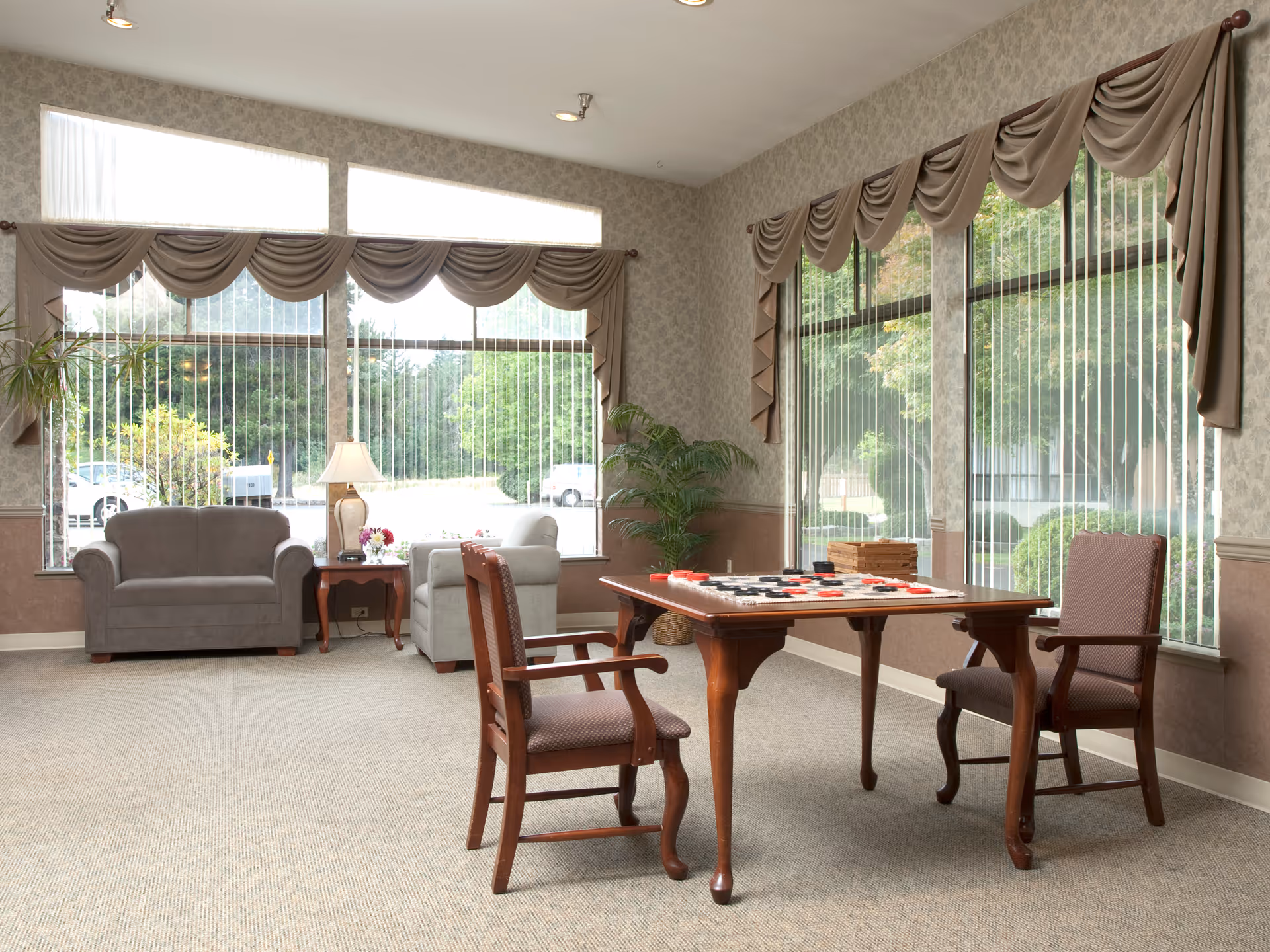 A bright and cozy sitting area with large windows covered by vertical blinds and decorative valances. The room features a wooden table with a checkers game set up, two upholstered chairs around the table, a gray loveseat, a gray armchair, a side table with a lamp and flowers, and a potted plant in the corner. Outside the windows, greenery and parked cars are visible.
