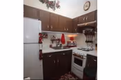 A small kitchen with dark brown cabinets, a white refrigerator, a white stove with an oven, and various kitchen utensils hanging on the wall. There are red accents including a towel and a rug, and a clock is visible on the wall above the cabinets.