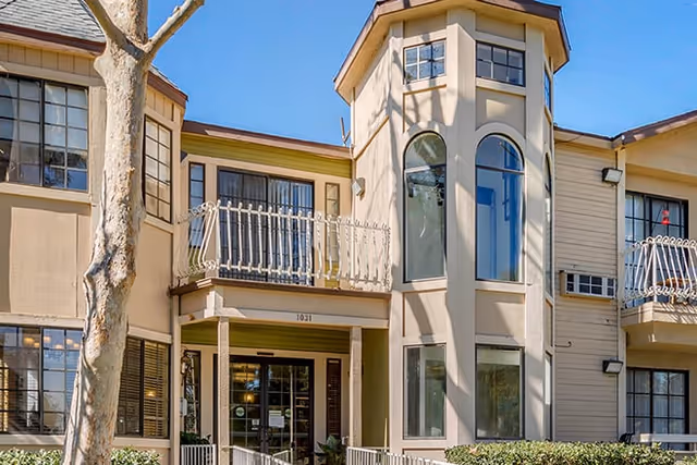 Exterior view of a two-story senior living facility building with beige walls, multiple windows, balconies with white railings, and a central entrance under a small porch. A tree is visible in front of the building under a clear blue sky.