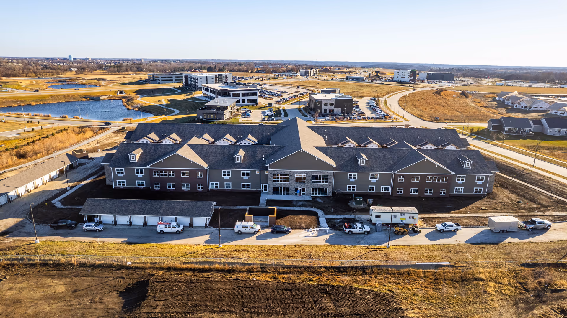 Aerial view of a large two-story senior living building with parked vehicles, surrounding roads, and nearby ponds and development.
