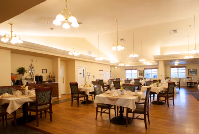 A spacious dining room with multiple tables covered in white tablecloths, each set with plates, glasses, and napkins. The room has wooden flooring, several chandeliers hanging from the ceiling, and large windows letting in natural light. Chairs with patterned upholstery surround the tables, and a sideboard with coffee and tea supplies is visible against one wall.