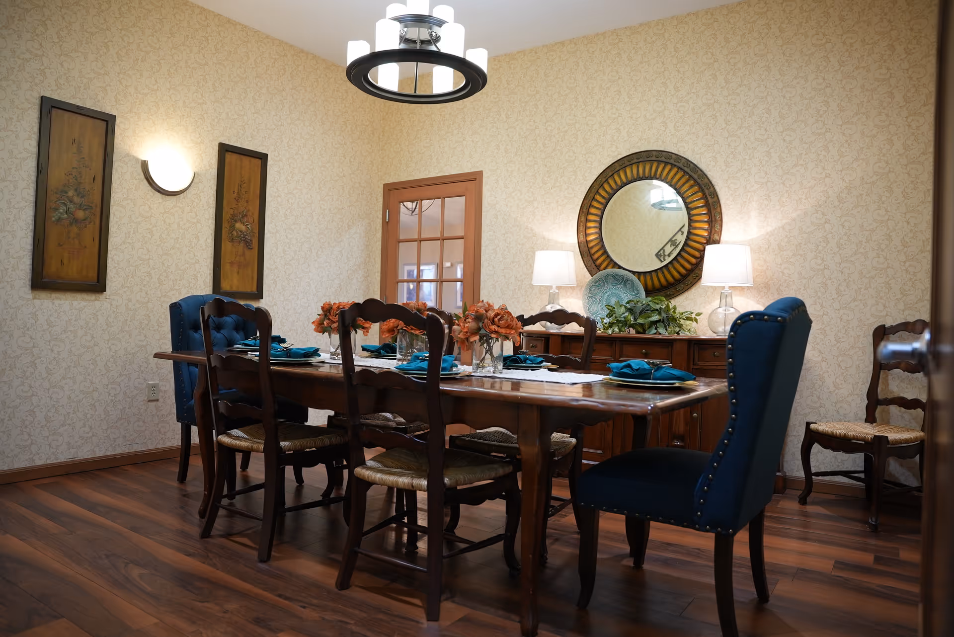 A dining room with a wooden table set for six people. The table has teal napkins, plates, and floral centerpieces. There are six chairs around the table, two of which are upholstered in blue fabric with nailhead trim. The room has wooden flooring, patterned wallpaper, a round decorative mirror on the wall, two table lamps, and framed artwork on the wall.