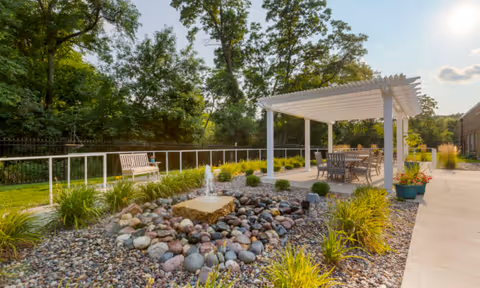 Sunlit outdoor courtyard with a white pergola dining area, a rock fountain, benches, and landscaped plants.