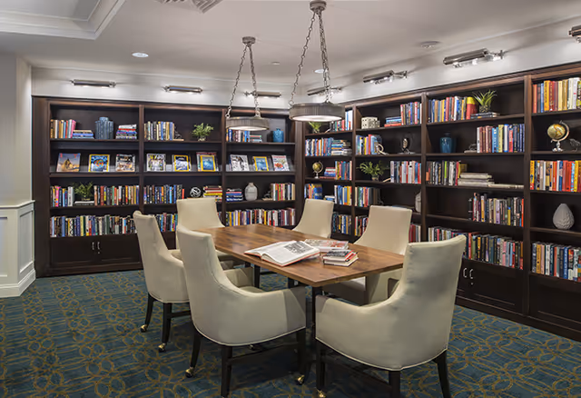 A cozy library room with dark wooden bookshelves filled with books, framed pictures, and decorative items. In the center, there is a wooden table surrounded by six cream-colored upholstered chairs. The room has a patterned carpet and is illuminated by ceiling lights and two hanging pendant lights above the table.