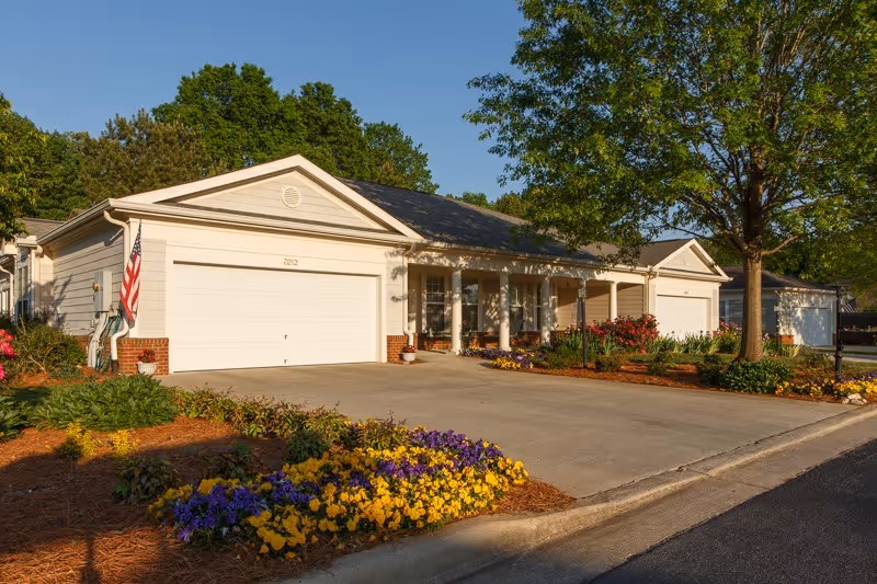 Exterior view of a single-story residential-style building with a white garage door, a porch with columns, and a driveway. The front yard has colorful flowers and a large tree, with a clear blue sky in the background.