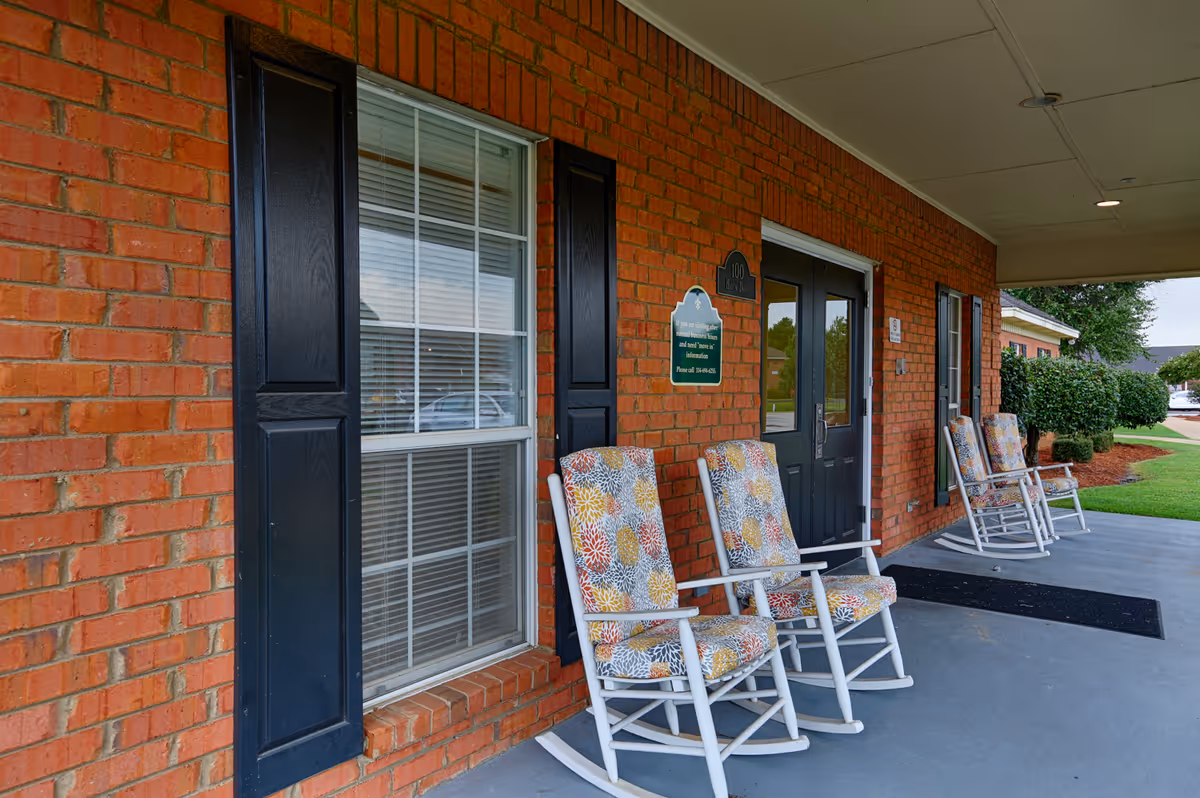 Covered porch area of a brick building with four white rocking chairs featuring colorful floral cushions. The porch has a concrete floor and black double doors with windows. There are black shutters on the windows and a green sign mounted on the wall next to the doors. Shrubs and a lawn are visible in the background.