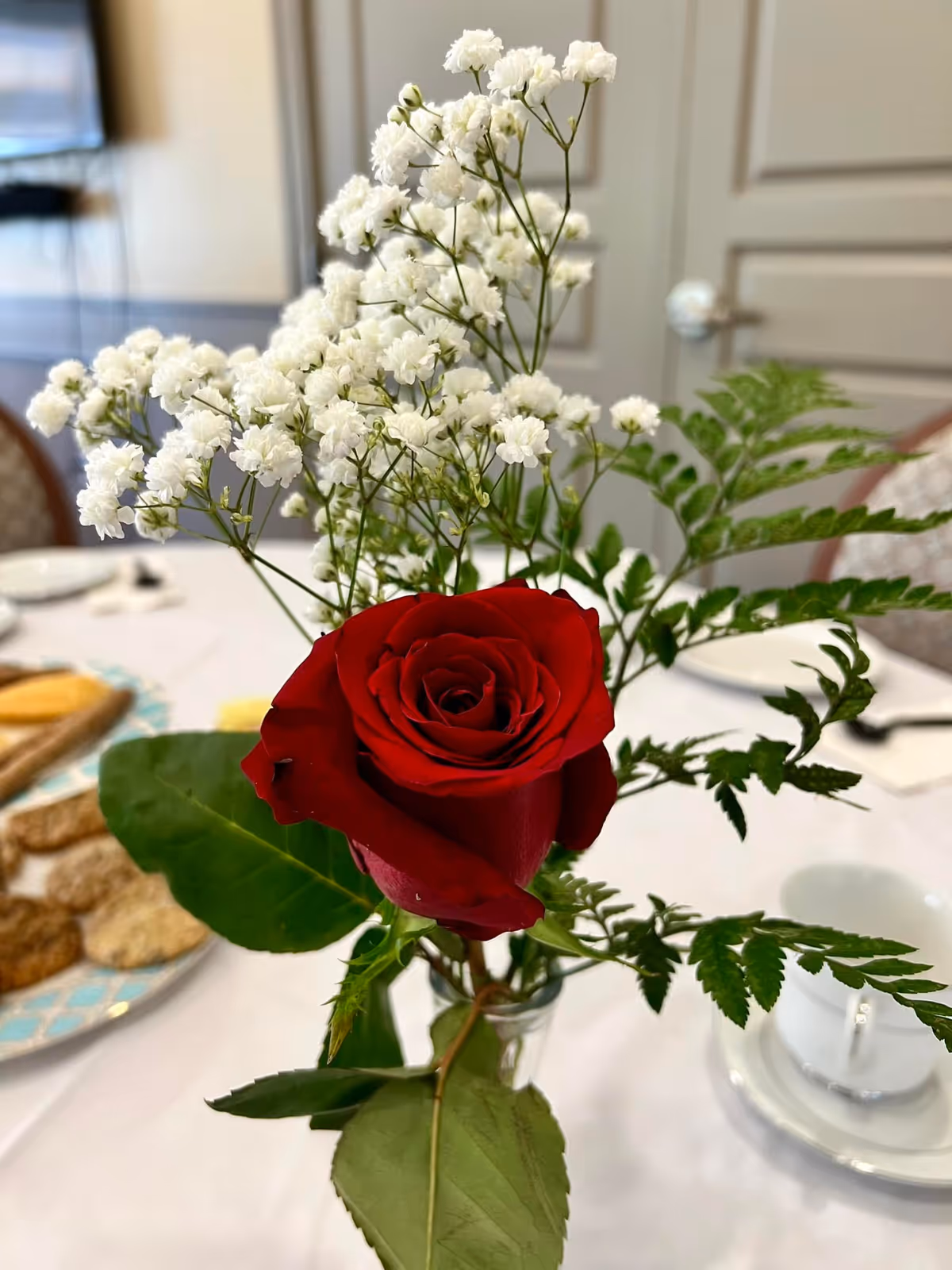 A red rose and white baby's breath arranged in a vase as a table centerpiece with plates and a teacup on a white tablecloth.