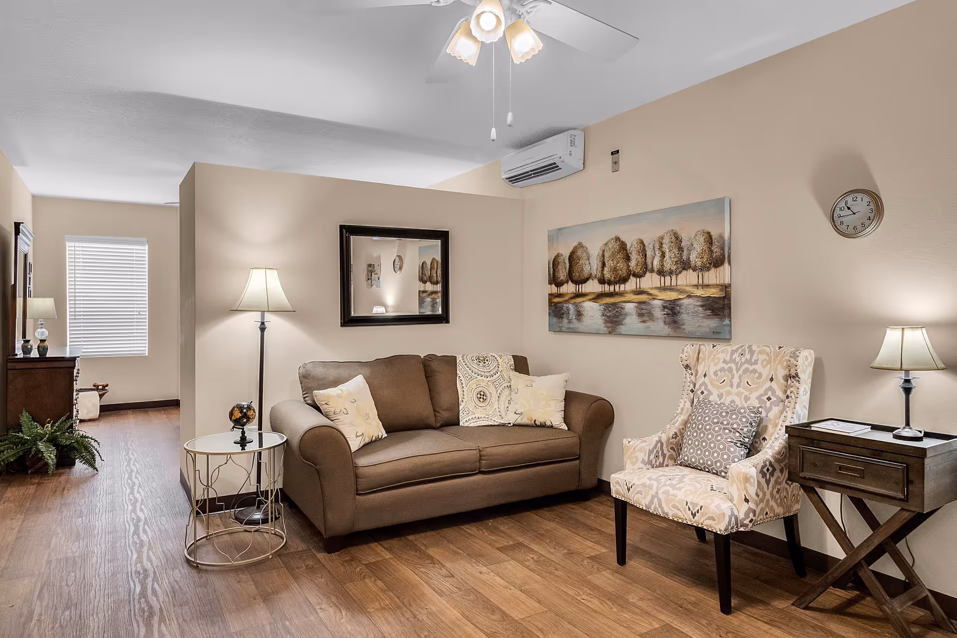 A cozy living room with a brown sofa adorned with decorative pillows, a patterned armchair with a cushion, two side tables each with a lamp, a wall clock, a ceiling fan with lights, and artwork depicting trees by a body of water. The room has wooden flooring and beige walls.