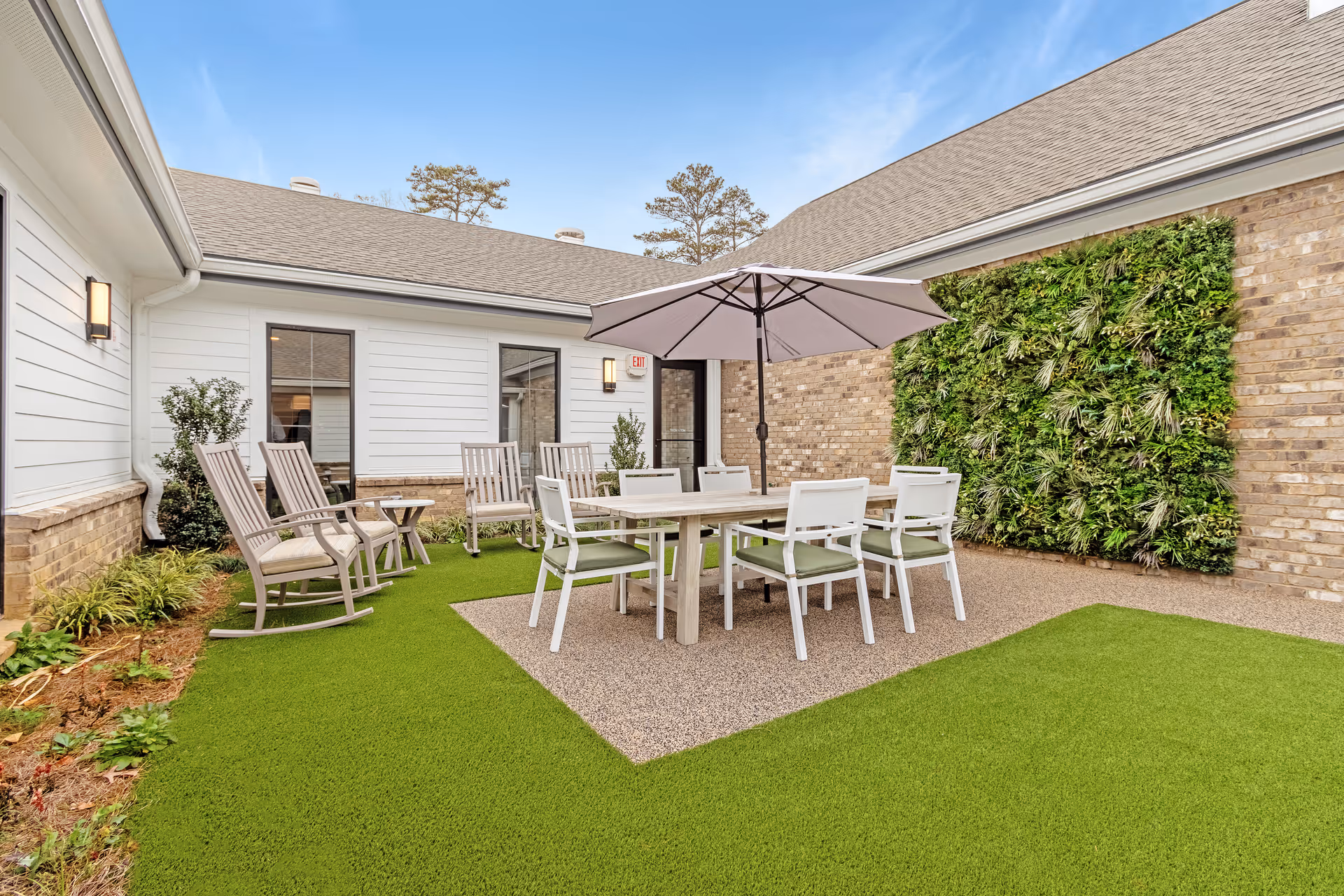 Outdoor patio area with a rectangular table surrounded by six white chairs with green cushions, shaded by a large umbrella. To the left, there are four wooden rocking chairs. The patio is surrounded by a green artificial lawn, a vertical garden on a brick wall, and white siding walls with windows.
