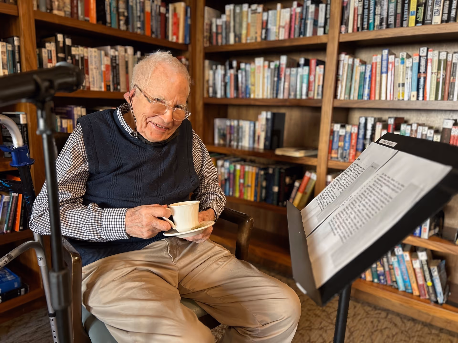 An elderly man smiling while holding a teacup and saucer in a book-lined room beside a music stand.