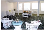 Sunlit communal living room with white wicker chairs and tables, floral cushions, a small dining set, and large windows.