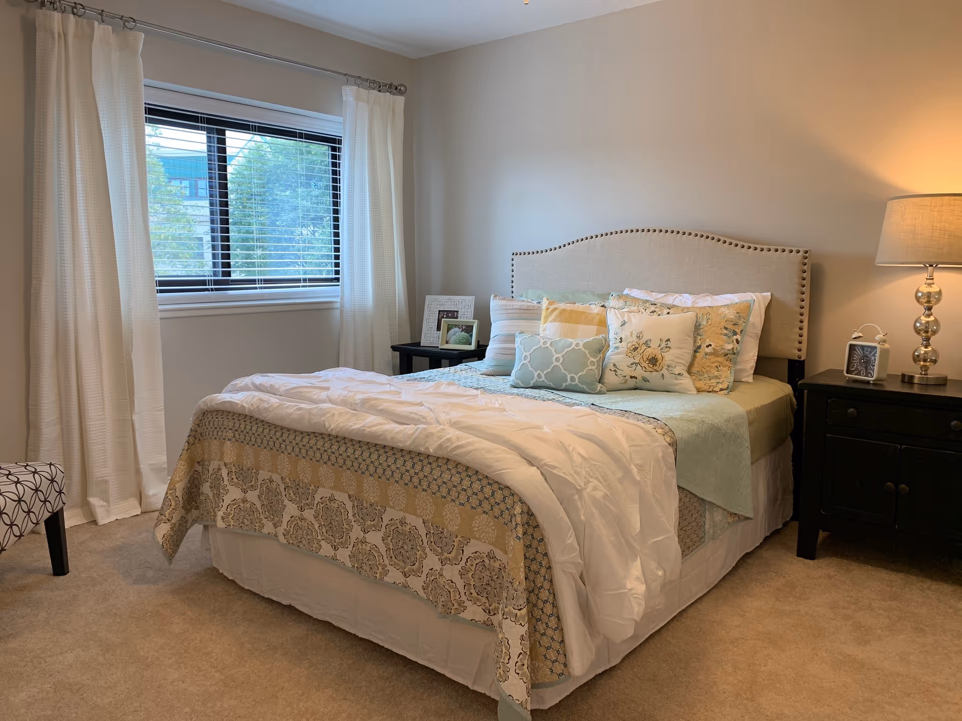 Bright, softly lit bedroom with a neatly made bed topped with decorative pillows, nightstands and a lamp beside a window with white curtains.