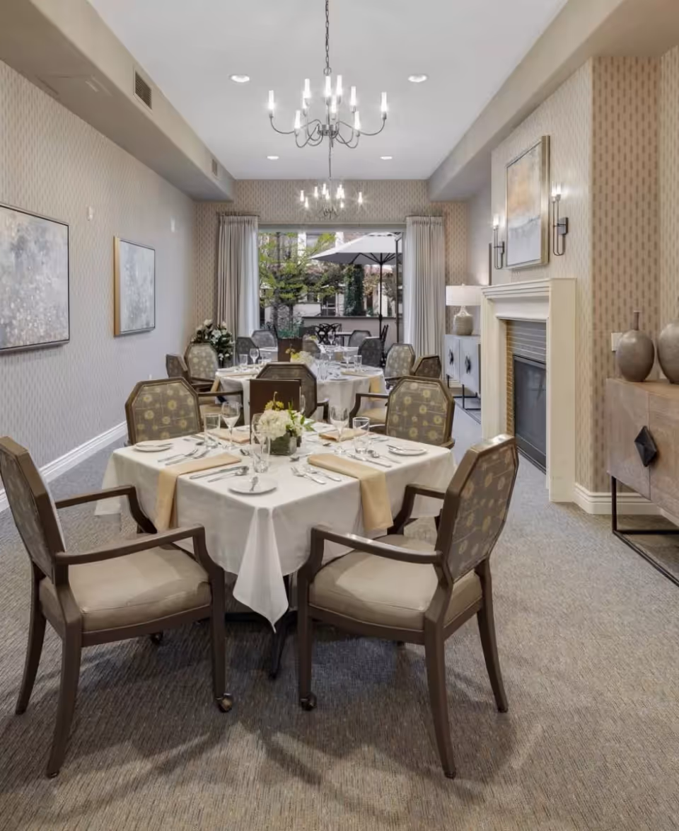A well-lit dining room with two round tables covered with white tablecloths and beige runners, each set with plates, glasses, and silverware. The room features upholstered chairs with patterned backs, a fireplace with a mantel, wall sconces, framed artwork, and a chandelier. Large windows at the back reveal an outdoor patio area with tables and umbrellas.