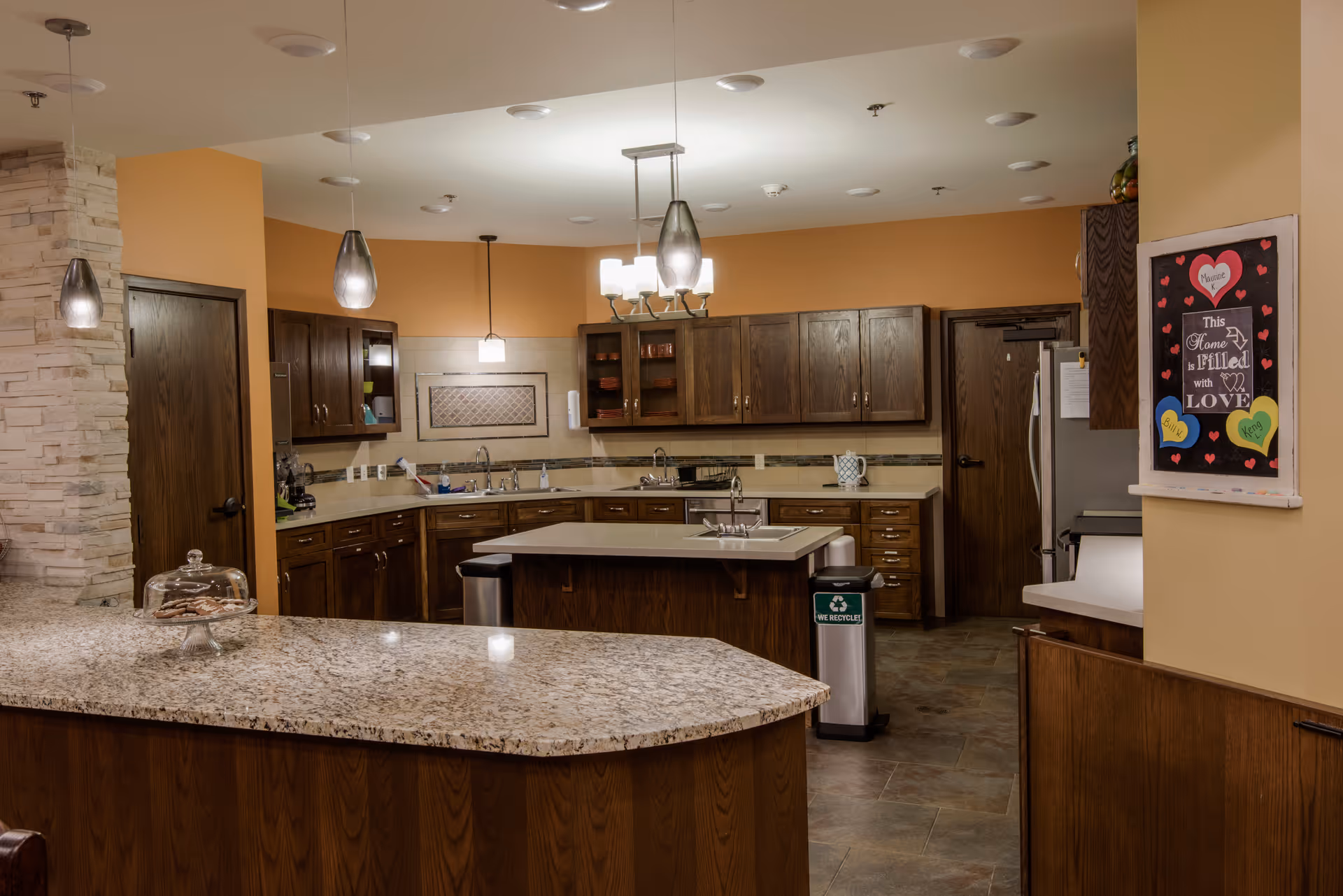 A spacious kitchen area with wooden cabinets, granite countertops, and modern pendant lighting. The kitchen features a central island with a sink, a recycling bin, and various kitchen appliances. There is a decorative sign on the wall that reads 'This Home is Filled with Love' surrounded by colorful heart shapes. The walls are painted in warm tones, and the floor is tiled.