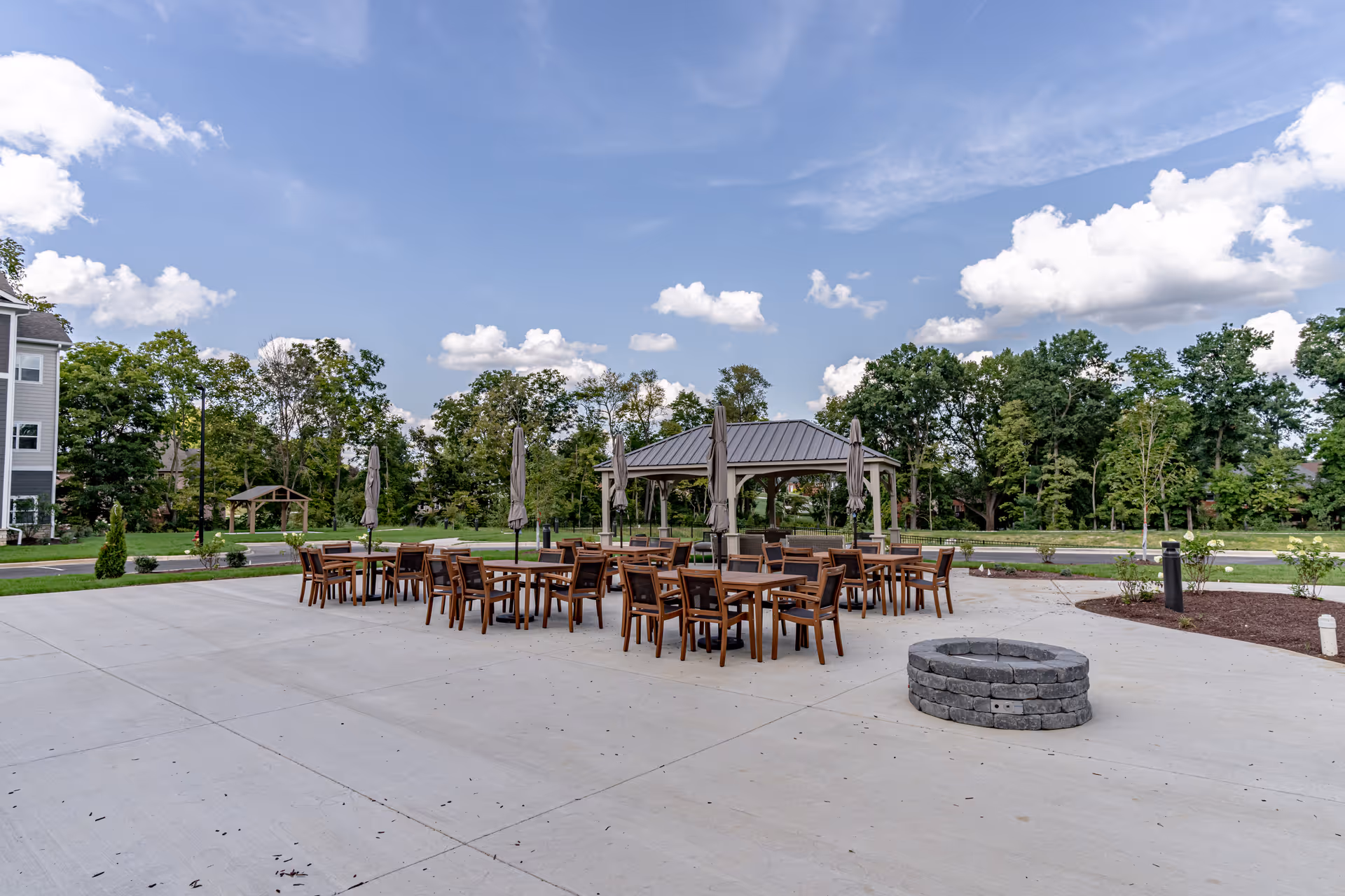 Outdoor patio area with multiple wooden tables and chairs arranged around a fire pit. There is a gazebo with a metal roof in the background, surrounded by trees and greenery under a partly cloudy blue sky.