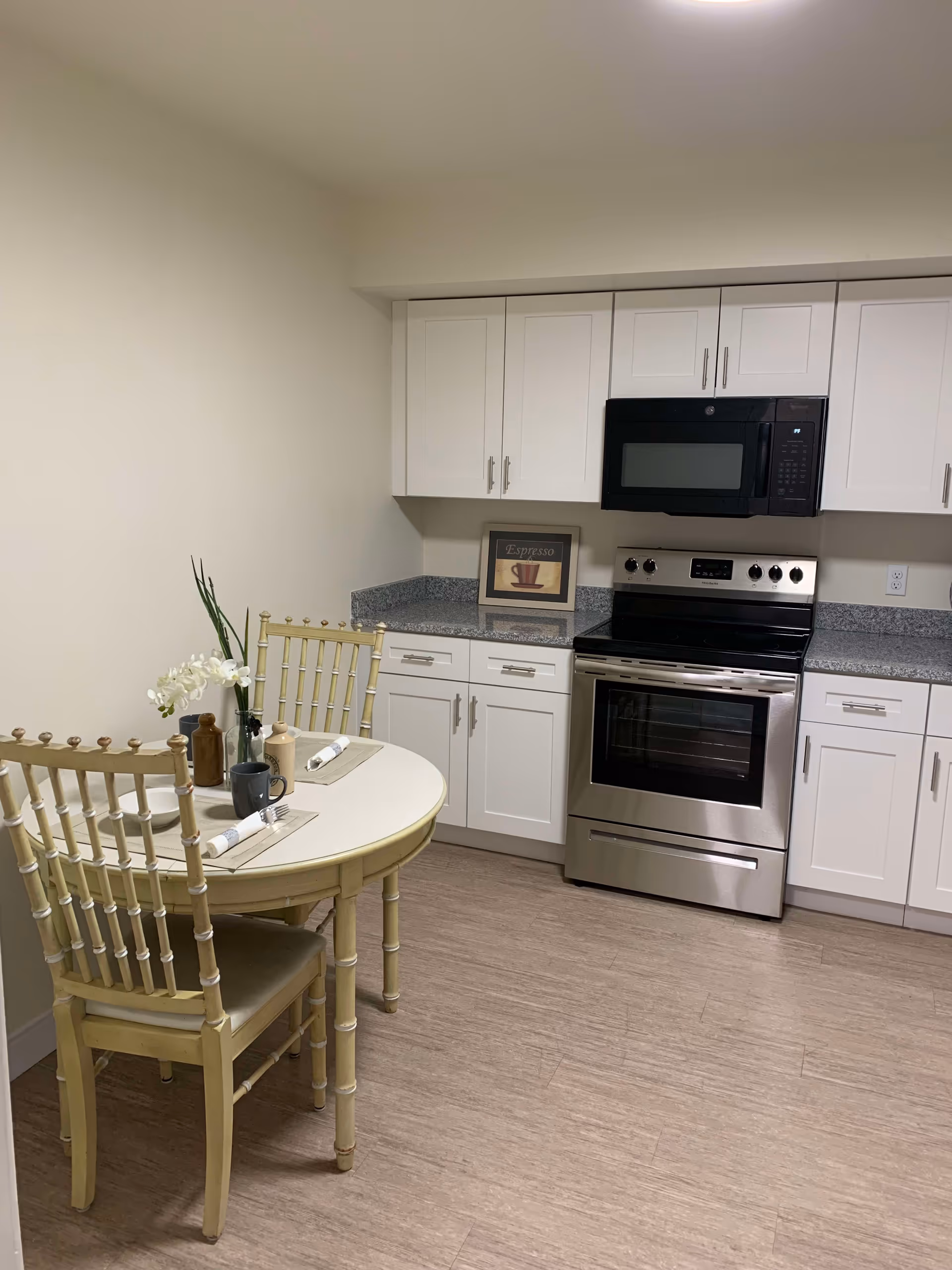 A small kitchen area with white cabinets, a stainless steel oven and microwave, and a round dining table with two wooden chairs. The table is set with two place settings, including napkins, forks, mugs, and a small vase with white flowers. There is a framed picture on the countertop with the word 'Espresso' and a coffee cup illustration.