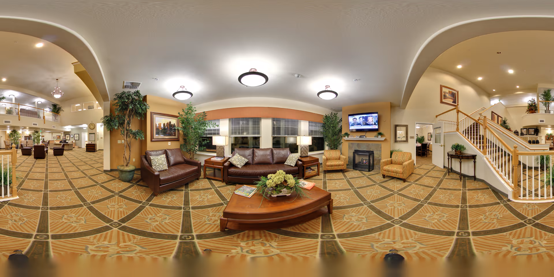 A spacious senior living facility common area with a patterned carpet, leather sofas, armchairs, a wooden coffee table with a floral centerpiece, and large windows with blinds. The room features warm lighting from ceiling fixtures and table lamps, a fireplace with a mounted TV above it, and decorative plants. There is a staircase with wooden railings leading to an upper floor, and additional seating areas are visible in the background.