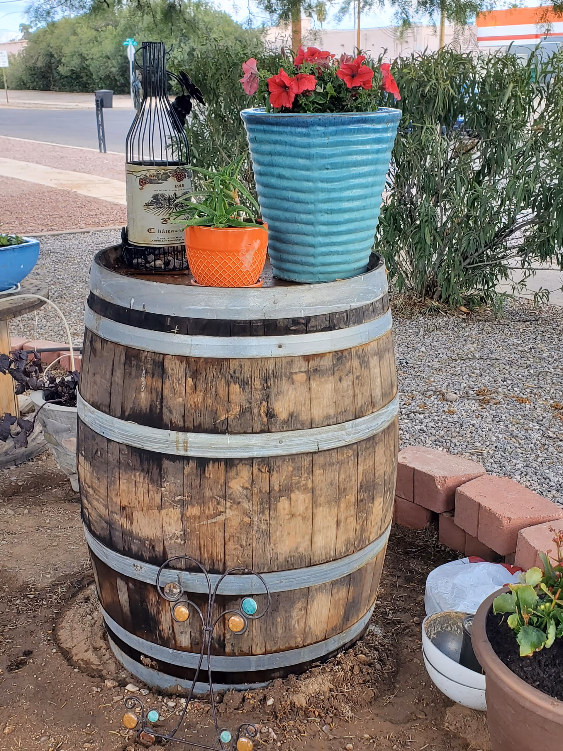 A rustic wooden barrel used as a table outdoors, with a large blue ceramic pot containing red flowers and a smaller orange pot with a green plant on top. There is also a decorative wine bottle holder shaped like a wine bottle. The barrel is placed on soil with some bricks and other potted plants nearby, and greenery and a street are visible in the background.