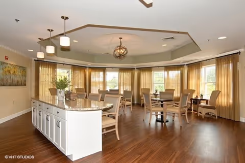 A bright dining room with large windows covered by sheer beige curtains. The room features multiple round tables with beige cushioned chairs and a long white island with a granite countertop in the foreground. Modern pendant lights hang from the ceiling, and there is a painting on the left wall. The floor is a warm wood tone.