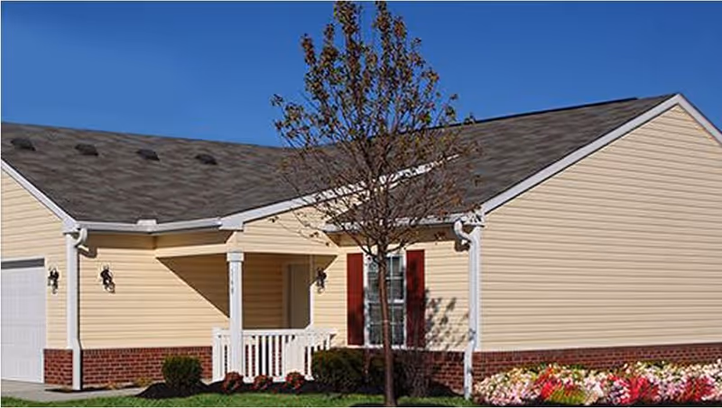Exterior view of a single-story senior living villa with beige siding, a gray shingled roof, a small front porch with white railing, a tree in the front yard, and colorful flowers along the foundation.