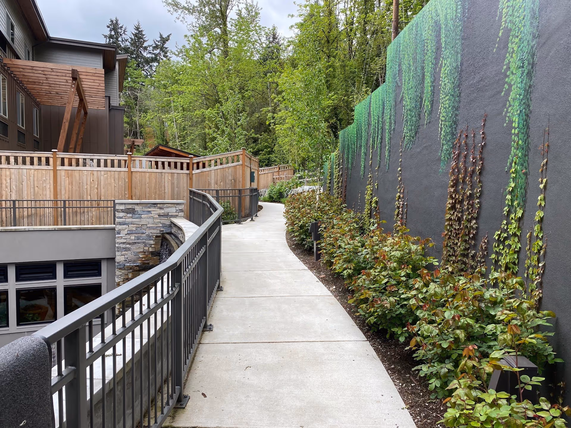 A paved outdoor walkway with metal railings on the left side and a tall wall on the right side covered with green climbing plants. There are bushes planted along the base of the wall and trees in the background. Part of a building with wooden fencing and stone accents is visible on the left.