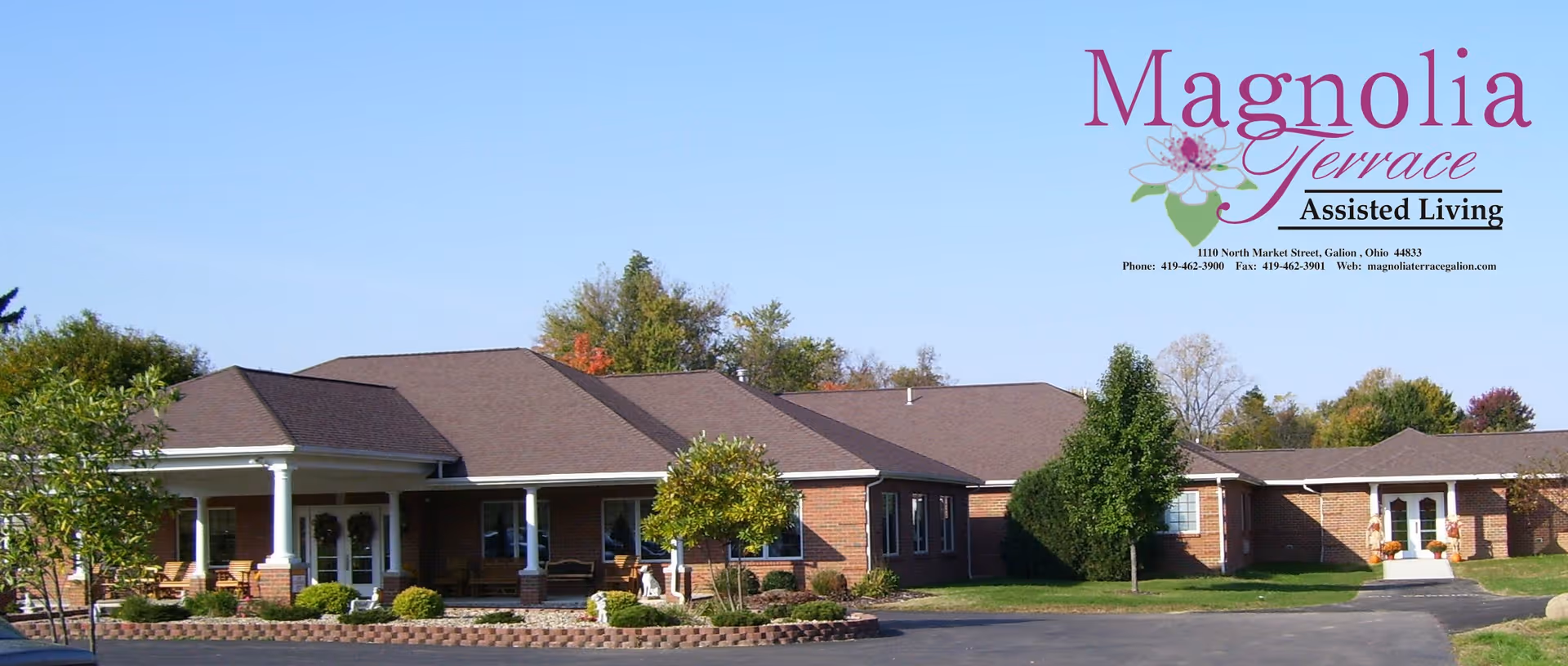 Exterior view of Magnolia Terrace Assisted Living facility showing a single-story brick building with a brown roof, surrounded by trees and shrubs, with a paved driveway in front.