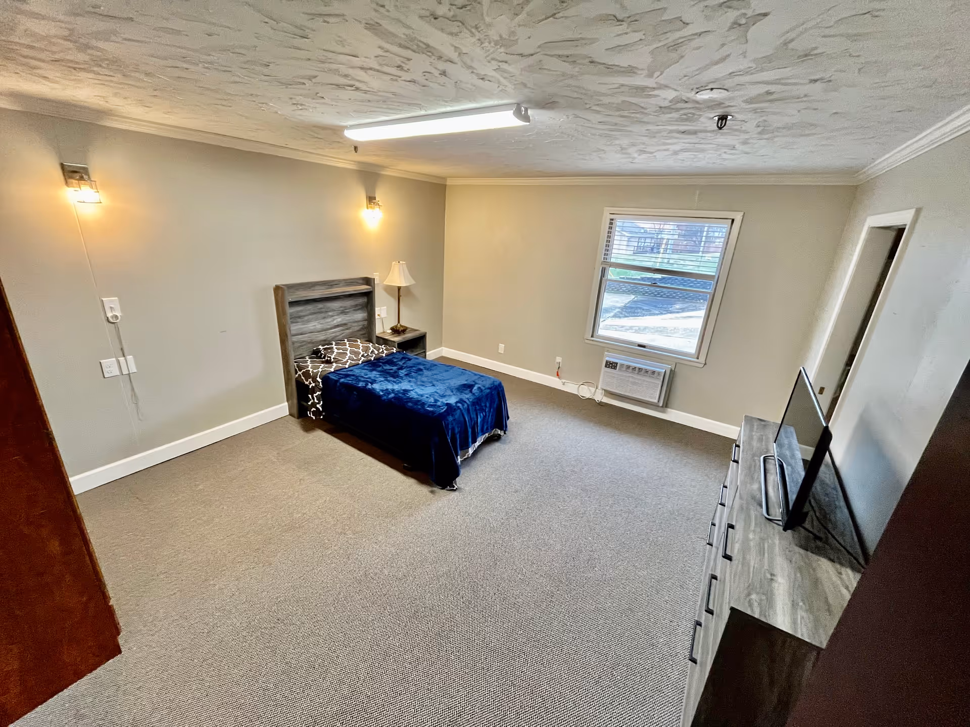 Furnished single-occupancy bedroom with a twin bed, nightstand and lamp, dresser with TV, window and wall-mounted air conditioner.