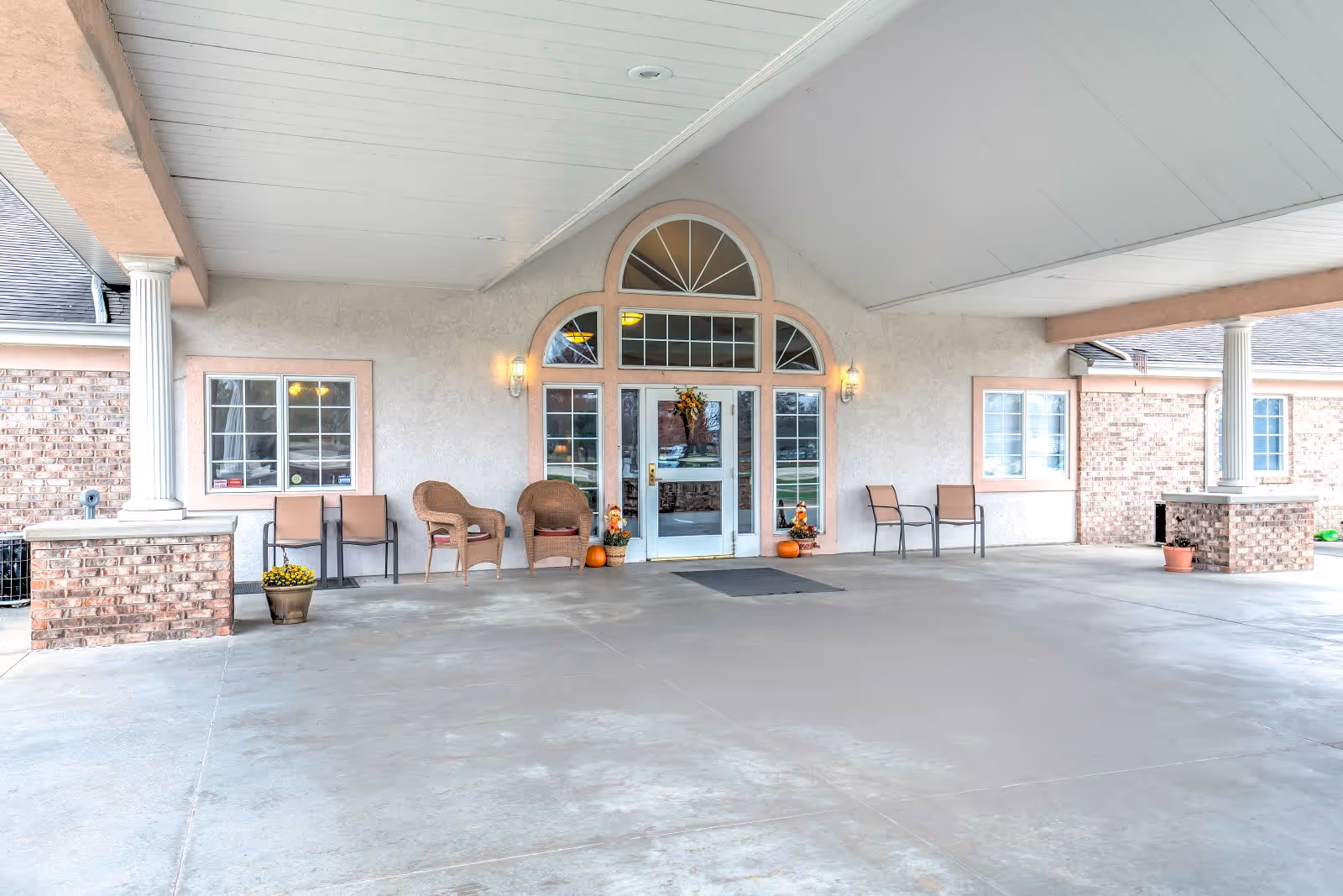 Covered entrance area of a building with a concrete floor, two white columns, several chairs arranged along the walls, potted plants, and a glass door with decorative windows above and on the sides. The entrance is decorated with autumn-themed wreaths and small pumpkins.