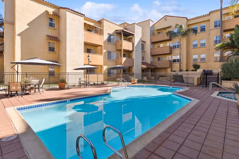Outdoor swimming pool area at The Remington Club with lounge chairs, tables with umbrellas, and a multi-story residential building in the background under a blue sky.