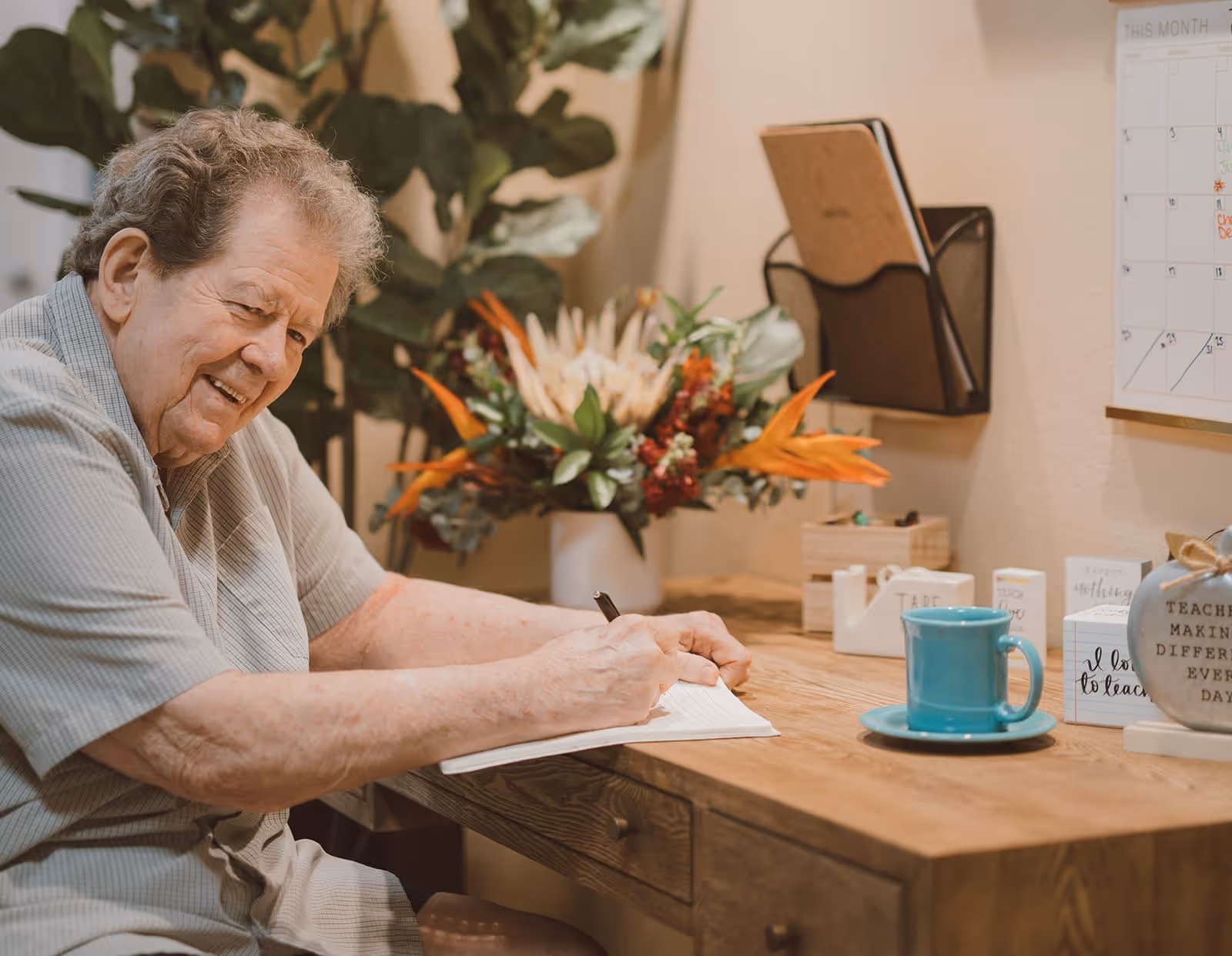 An elderly man sitting at a wooden desk writing in a notebook. The desk has a blue cup and saucer, decorative blocks with inspirational quotes, and a vase with colorful flowers. A calendar and wall organizer are mounted on the wall behind the desk.