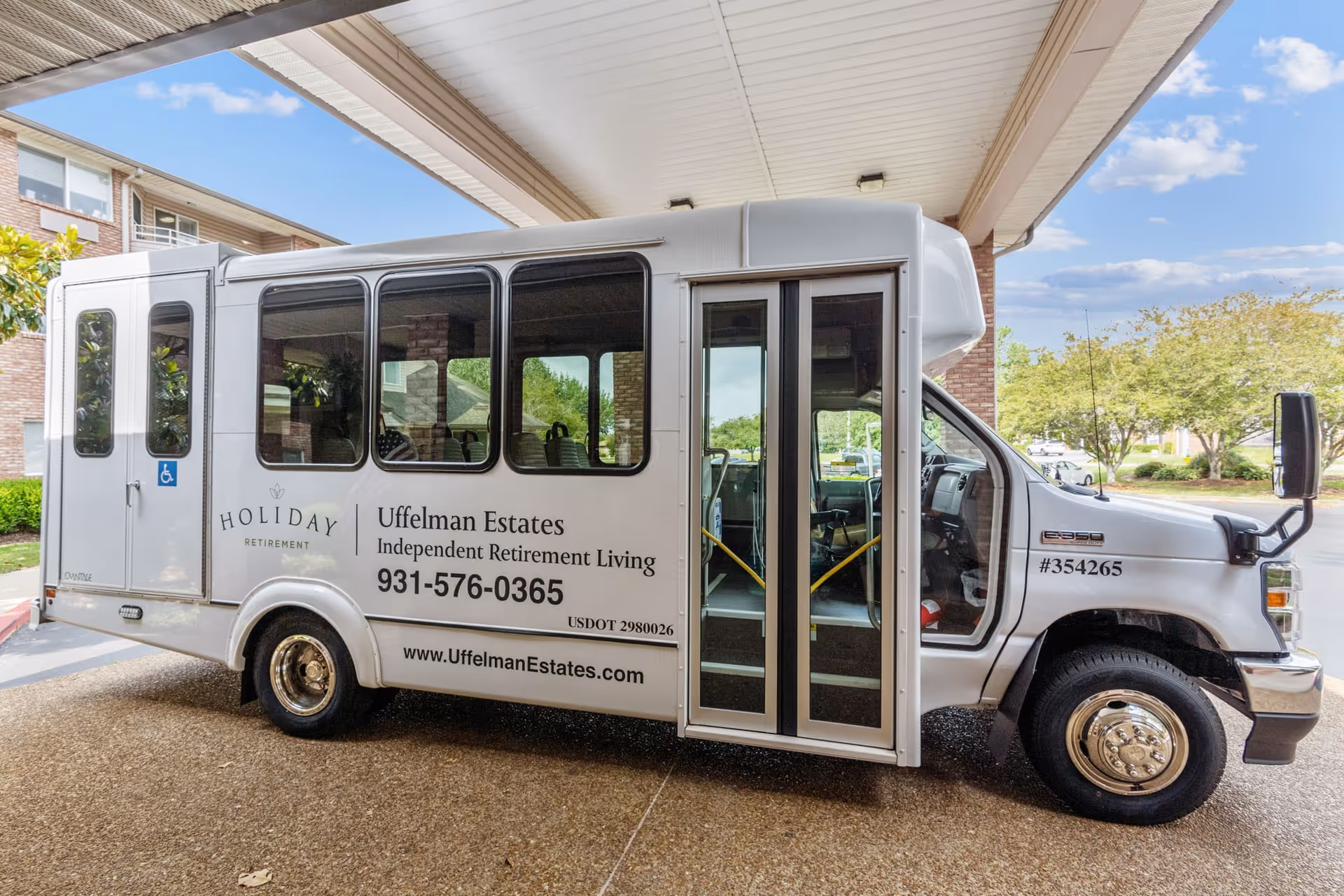 A white shuttle bus parked under a covered drop-off area at Holiday Uffelman Estates, an independent retirement living facility. The bus has large windows, a wheelchair accessibility sign, and text displaying the facility's name, phone number, and website.