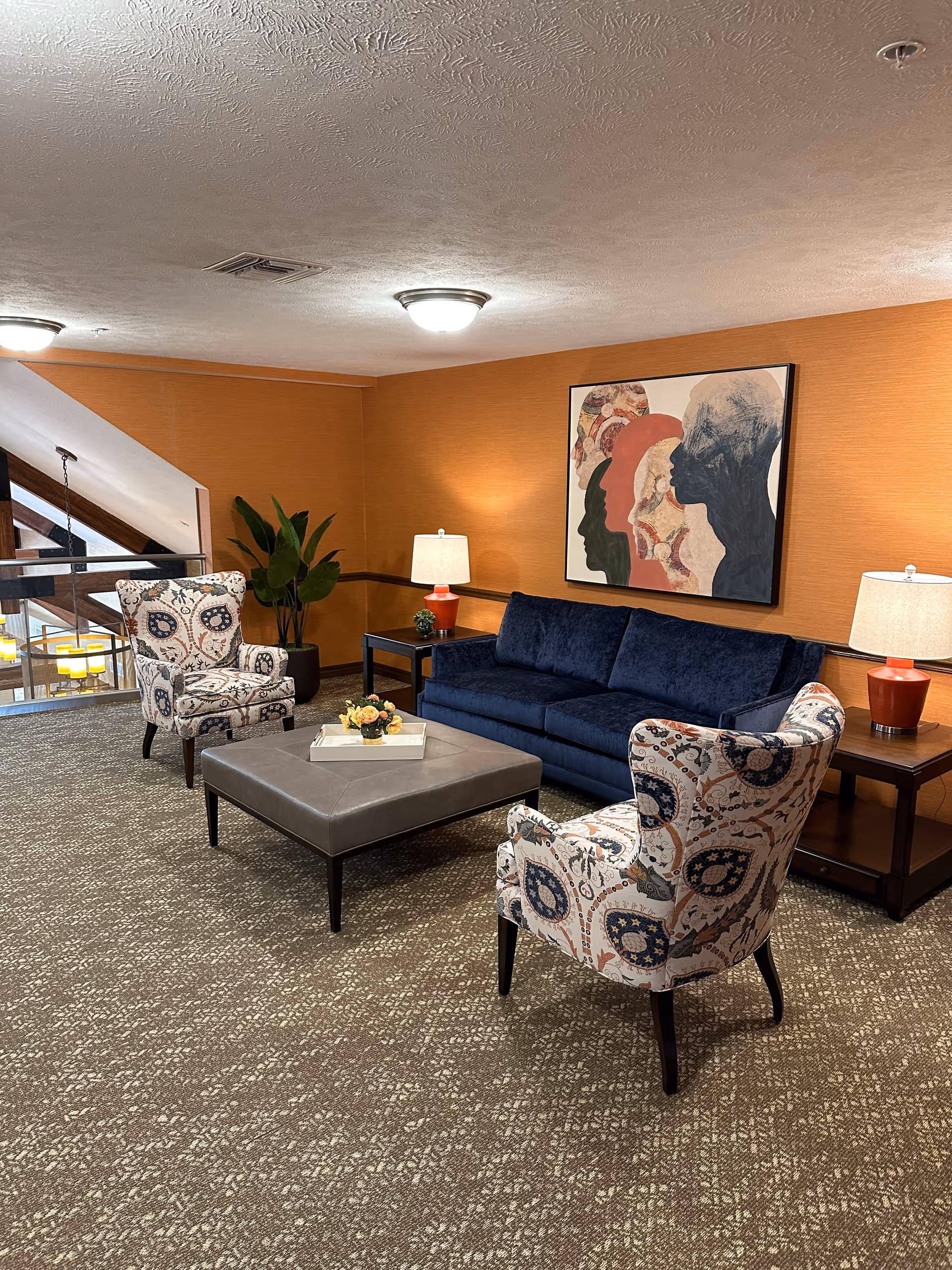 Lounge seating area with a navy sofa, two patterned armchairs, a gray ottoman coffee table, side tables with lamps, and artwork on an orange wall.