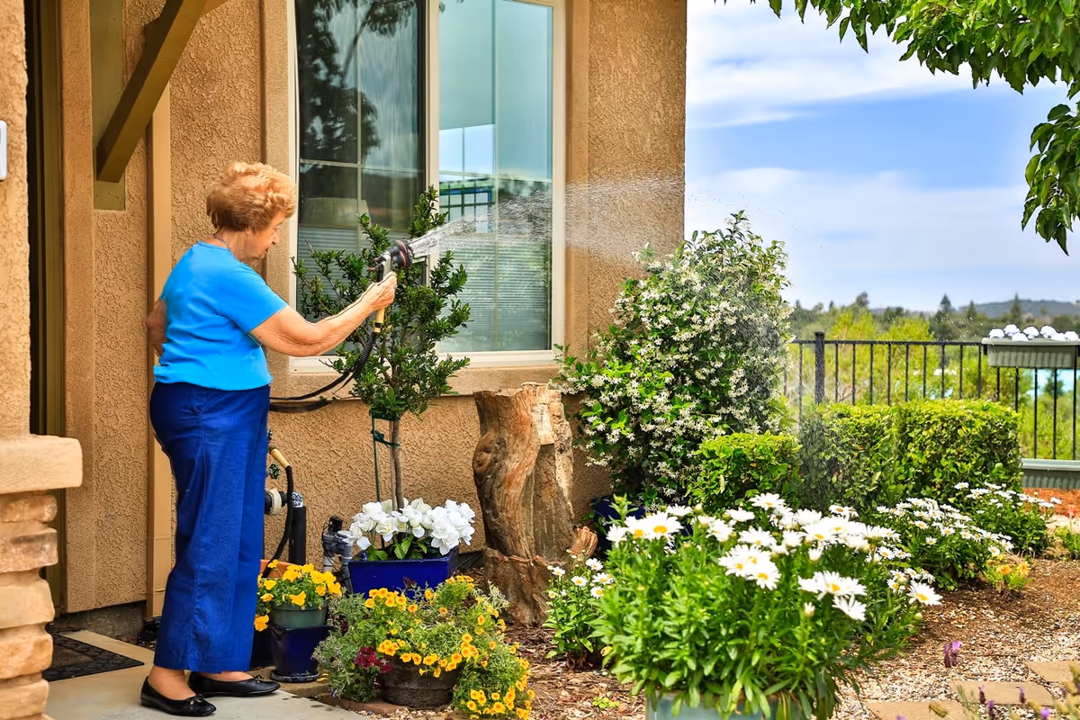 An elderly woman wearing a blue shirt and dark blue pants is watering plants with a garden hose outside a beige stucco building. The garden area is filled with various flowering plants and greenery, and there is a metal railing and trees in the background under a partly cloudy sky.