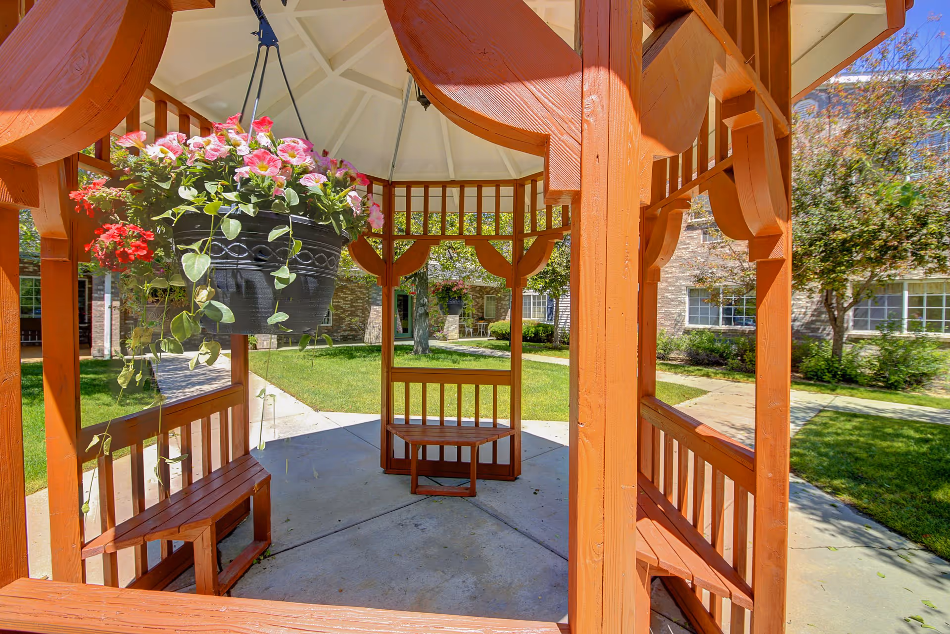 View from inside a wooden gazebo with benches, overlooking a green lawn and trees in an outdoor courtyard area of a senior living facility. A hanging flower pot with pink and red flowers is suspended from the gazebo roof.