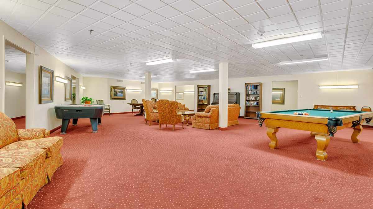 A spacious common room with red carpet flooring and white tiled ceiling. The room features a pool table with balls set up, an air hockey table, several upholstered chairs and sofas with a patterned orange fabric, wooden bookshelves filled with books, framed paintings on the walls, and a few tables and chairs in the background.