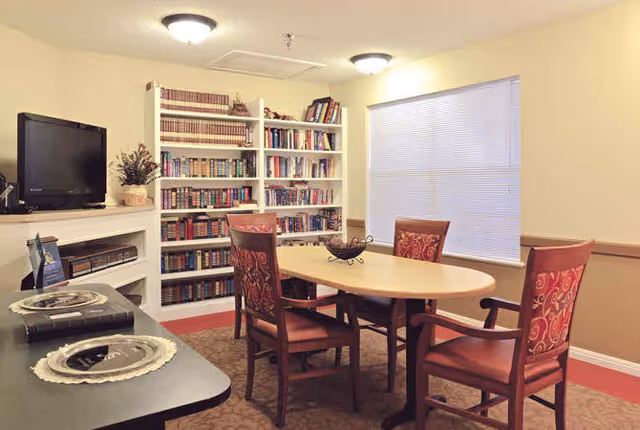 A cozy room with a round wooden table surrounded by four upholstered chairs with red patterned fabric. Behind the table is a large bookshelf filled with books. To the left, there is a flat-screen TV on a white mantel with decorative items. The room has beige walls, carpeted floor, and a window with closed blinds.