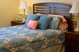 A neatly made bed with a wooden headboard, adorned with patterned blue and green bedding and several pillows in blue and coral colors. On each side of the bed are wooden nightstands with white lamps and a framed photo on the left nightstand. The walls are painted a soft yellow.