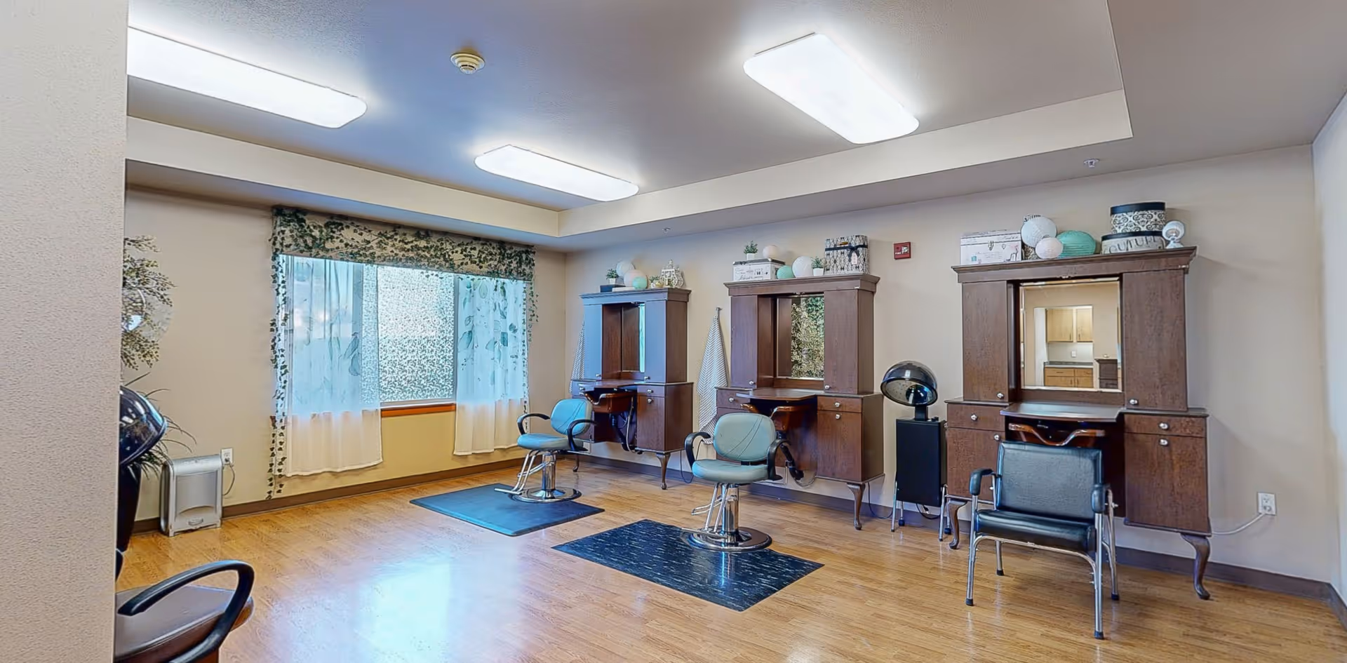 Interior of a salon area in a senior living facility with three styling stations, each with a mirror, wooden cabinetry, and salon chairs. The room has wood flooring, a large window with floral curtains, and bright overhead lighting.