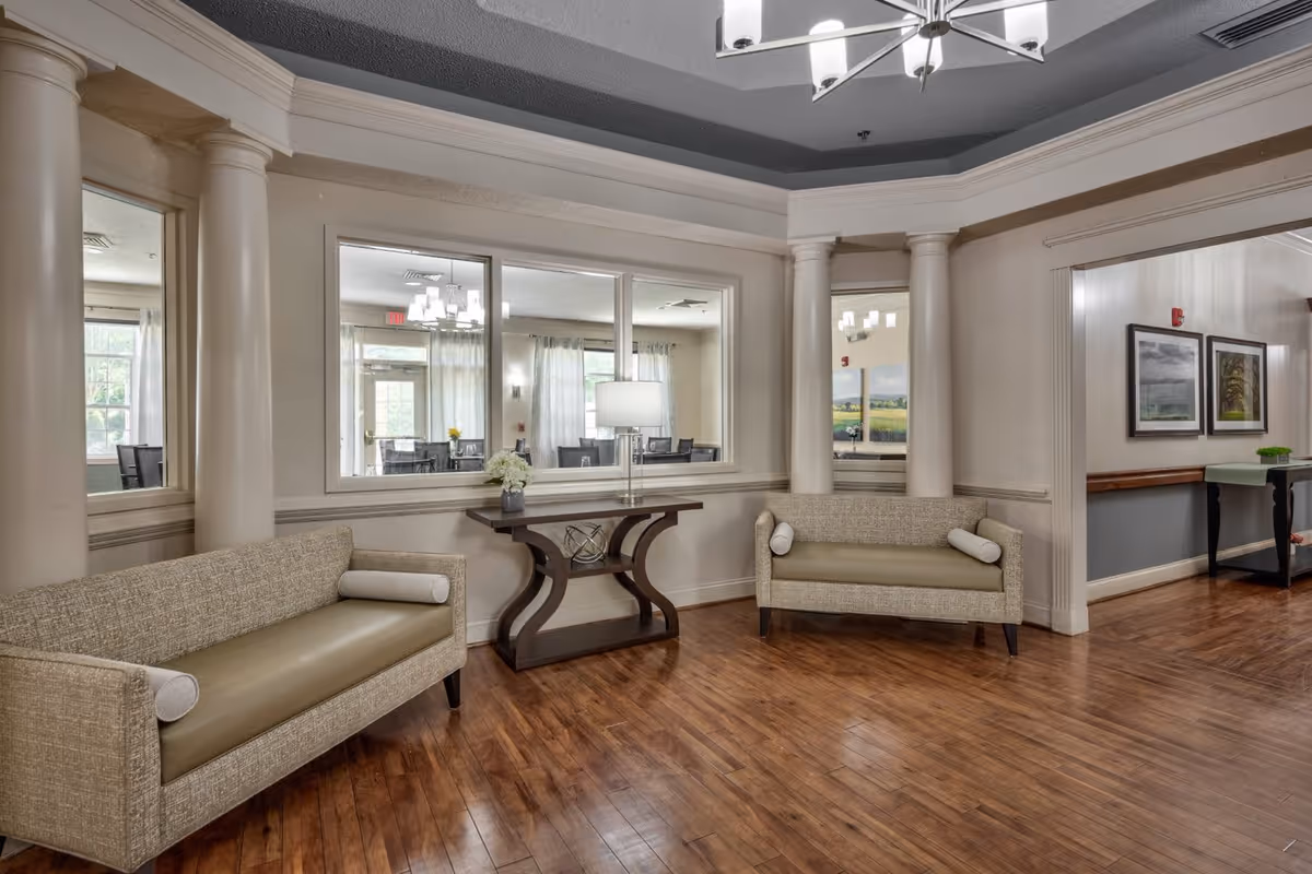 A spacious interior common area with two beige upholstered sofas with cylindrical pillows, a wooden table with a decorative object and a lamp, large windows showing a dining area with tables and chairs, wooden flooring, and white columns supporting the ceiling.