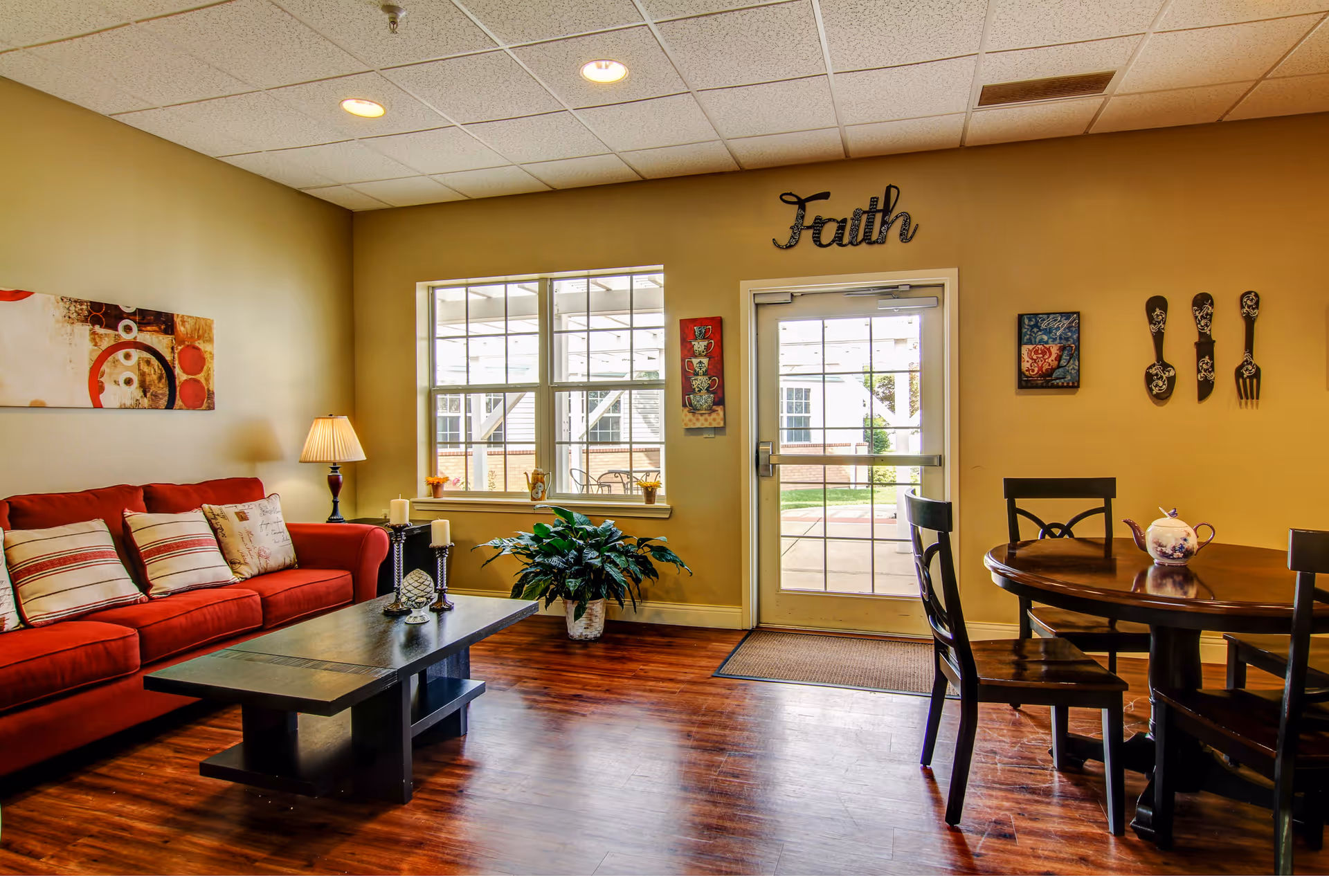 Bright communal living room with a red sofa, coffee table, dining table, plants, and a glass door beneath a 'Faith' wall sign.