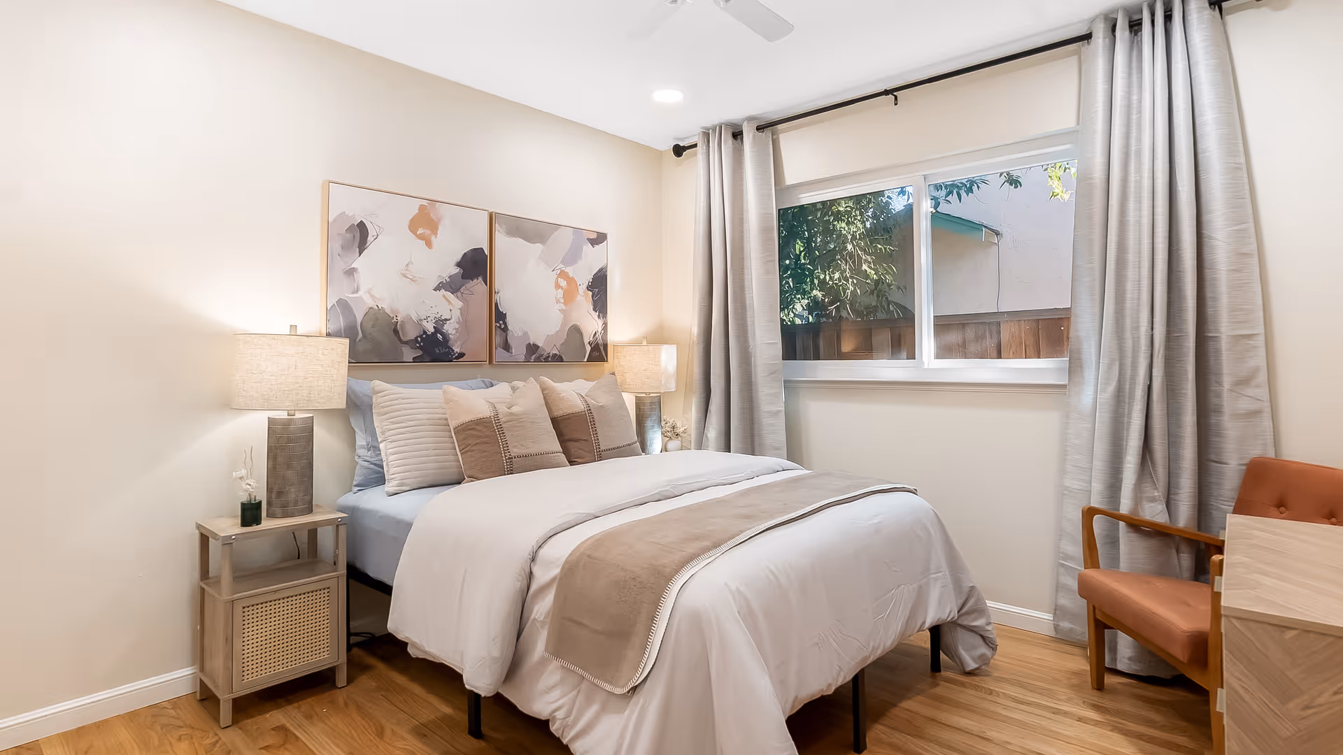 A bright, cozy bedroom with a made bed, bedside tables and lamps, artwork above the headboard and a window with curtains.