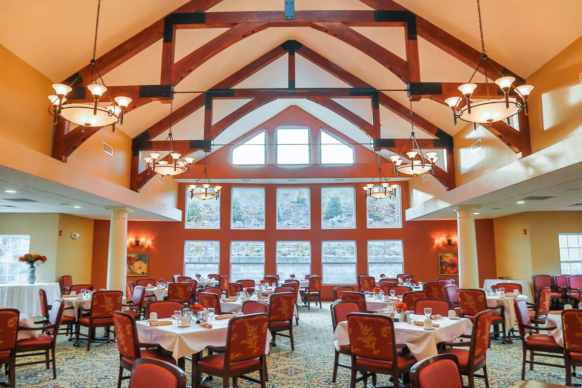 Spacious dining room with multiple tables covered in white tablecloths, each set with glasses, napkins, and small flower arrangements. The room features high vaulted ceilings with exposed wooden beams and several hanging chandeliers. Large windows on one wall let in natural light and offer a view of an outdoor garden area.