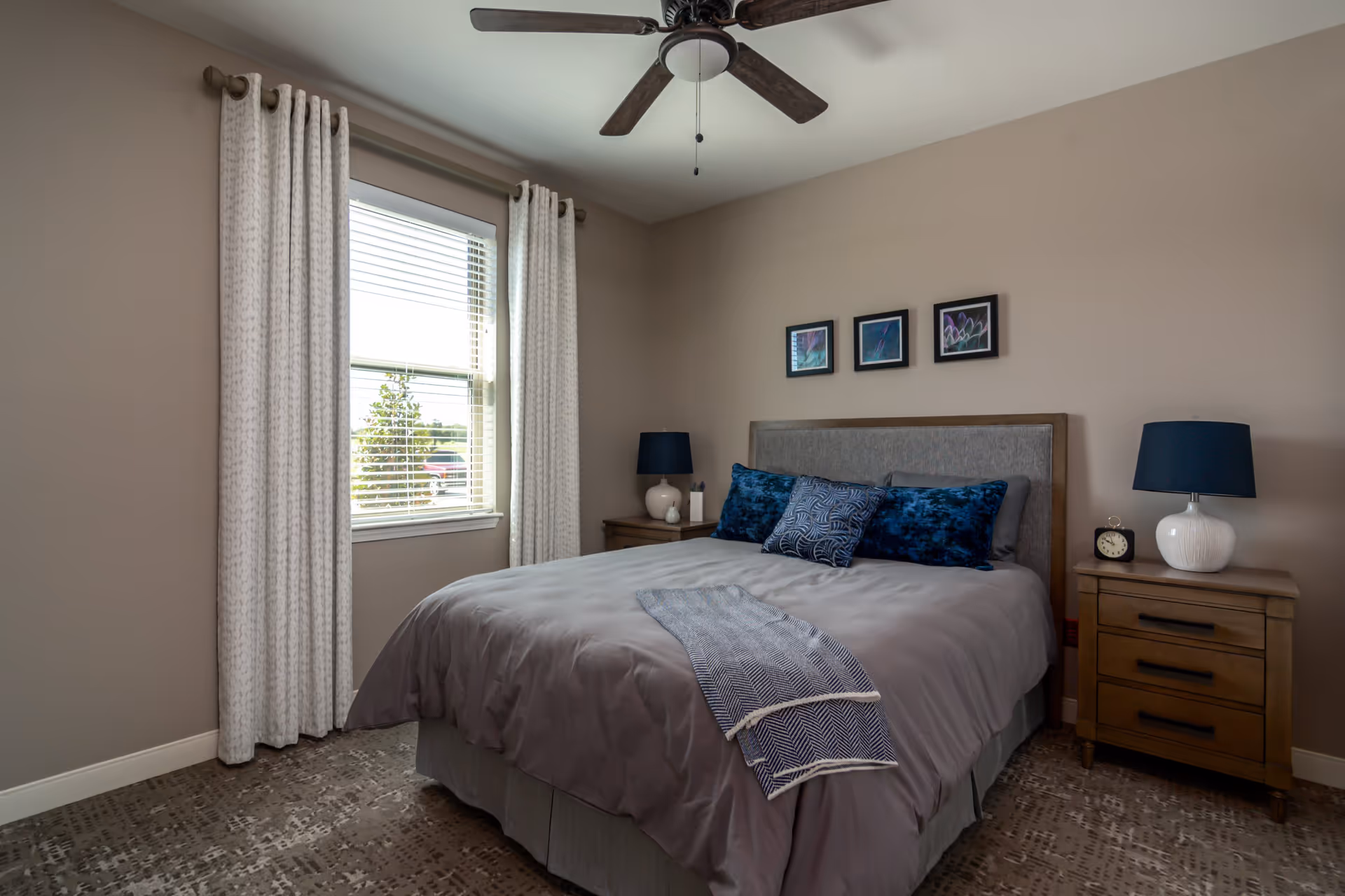 A neatly made bed with gray bedding and blue decorative pillows in a bedroom. There are two wooden nightstands on either side of the bed, each with a white lamp with a dark blue shade. A small clock is on the right nightstand. Above the bed are three framed pictures. A window with white curtains is on the left wall, and a ceiling fan with dark blades is mounted on the ceiling.