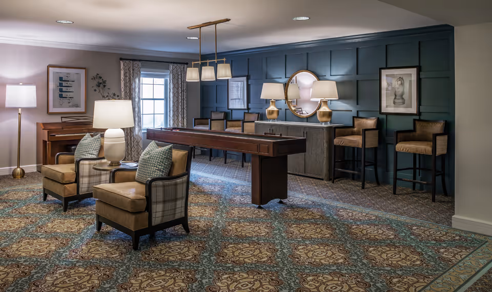 Well-appointed interior common room with upholstered chairs, a shuffleboard table, piano, and bar seating against a paneled accent wall.