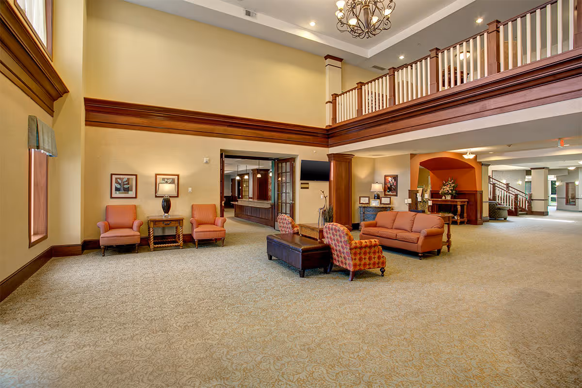 Spacious senior living facility common area with high ceilings, beige walls, and carpeted floor. The room features a seating arrangement with two orange armchairs and a small table against one wall, and a sofa with two patterned armchairs around a coffee table in the center. There is a chandelier hanging from the ceiling and a balcony railing overlooking the area. The space is well-lit with recessed lighting and has decorative wood trim and columns.